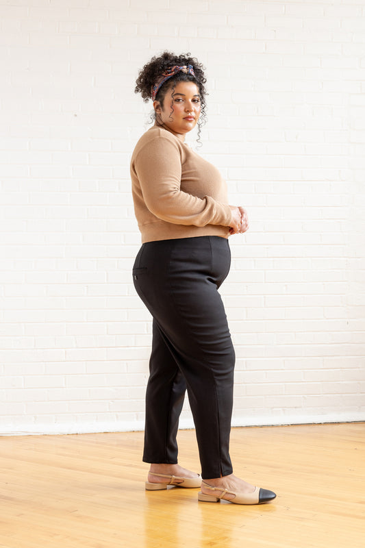 Person wearing a beige sweater and black pants standing against a white wall.