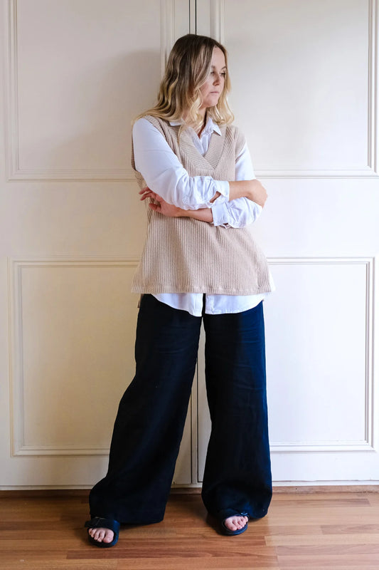 Woman wearing a beige vest over a white shirt with navy pants against a neutral wall.