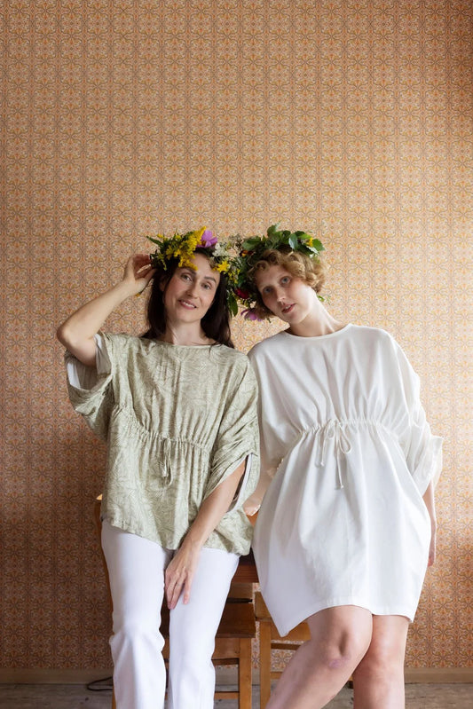 Two women wearing loose-fitting tops and white pants with floral crowns against a beige wall.