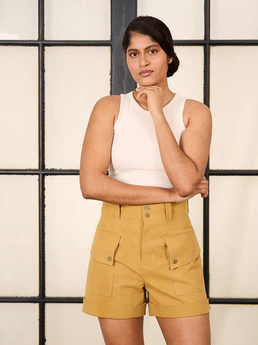 Woman wearing a white sleeveless top and mustard yellow shorts against a neutral background