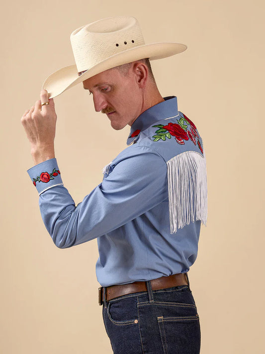 Man wearing a blue embroidered shirt with fringes and a cowboy hat on a beige background
