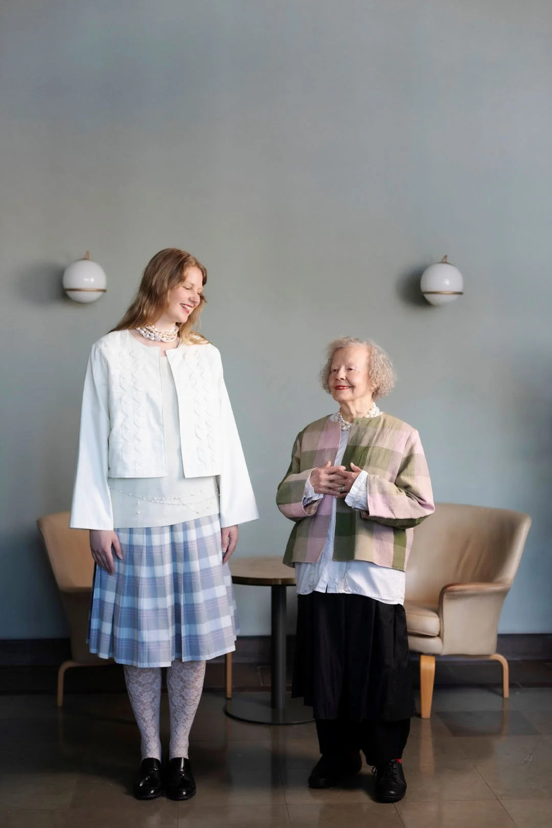 Two women standing in a room with a plain wall and chairs.