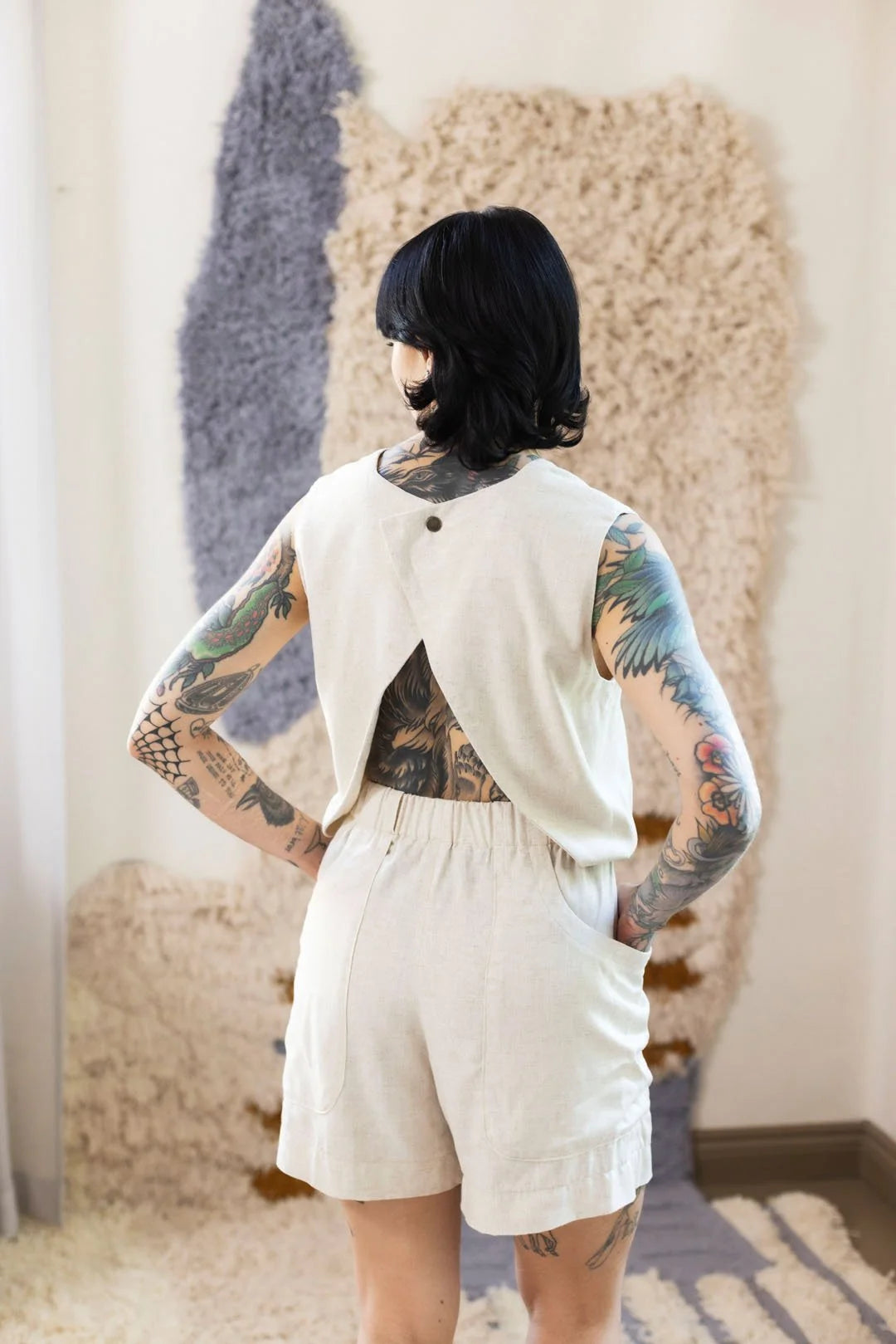 Person wearing a white outfit with tattoos visible on arms and back, standing in a room with textured wall art.