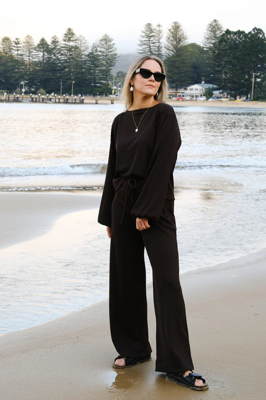 Woman in black outfit standing on a beach with trees and water in the background