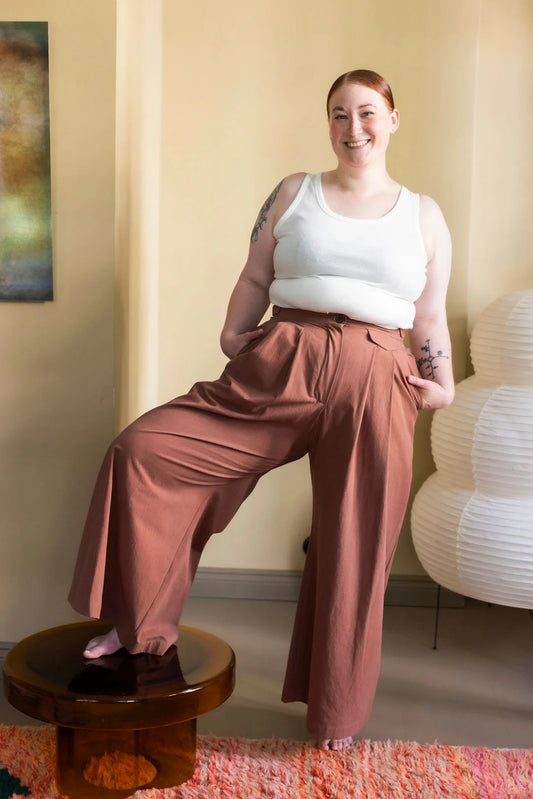 Woman wearing a white tank top and brown pants sitting on a small round stool in a room with a lamp and abstract painting.