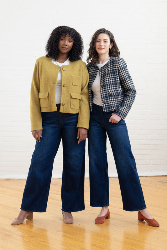 Two women standing side by side wearing mustard and plaid jackets with blue jeans against a white wall.