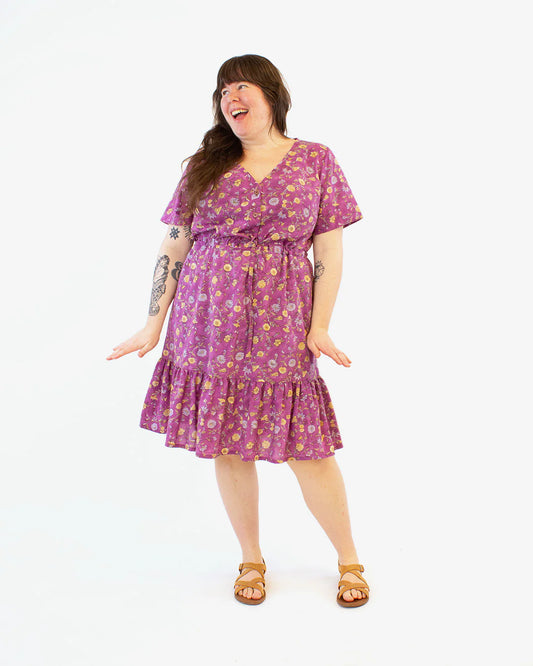 Full detail photo of woman wearing a pink floral print dress at midi-length with a front button placket, drawstring waist and a bottom tier. Photo taken against a white background.