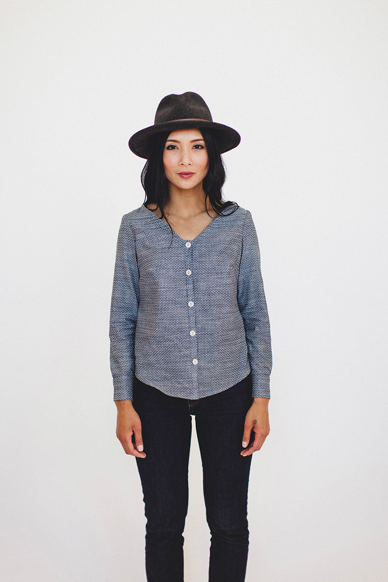Woman wearing a black hat, blue patterned shirt, and black pants on a white background