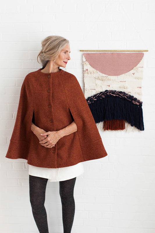 Woman wearing a brown cape standing in front of a wall with a textured piece of art.