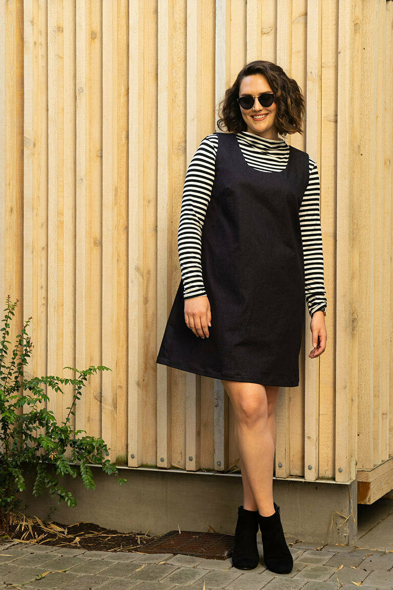 Woman wearing a black pinafore dress over a striped shirt against a wooden wall.