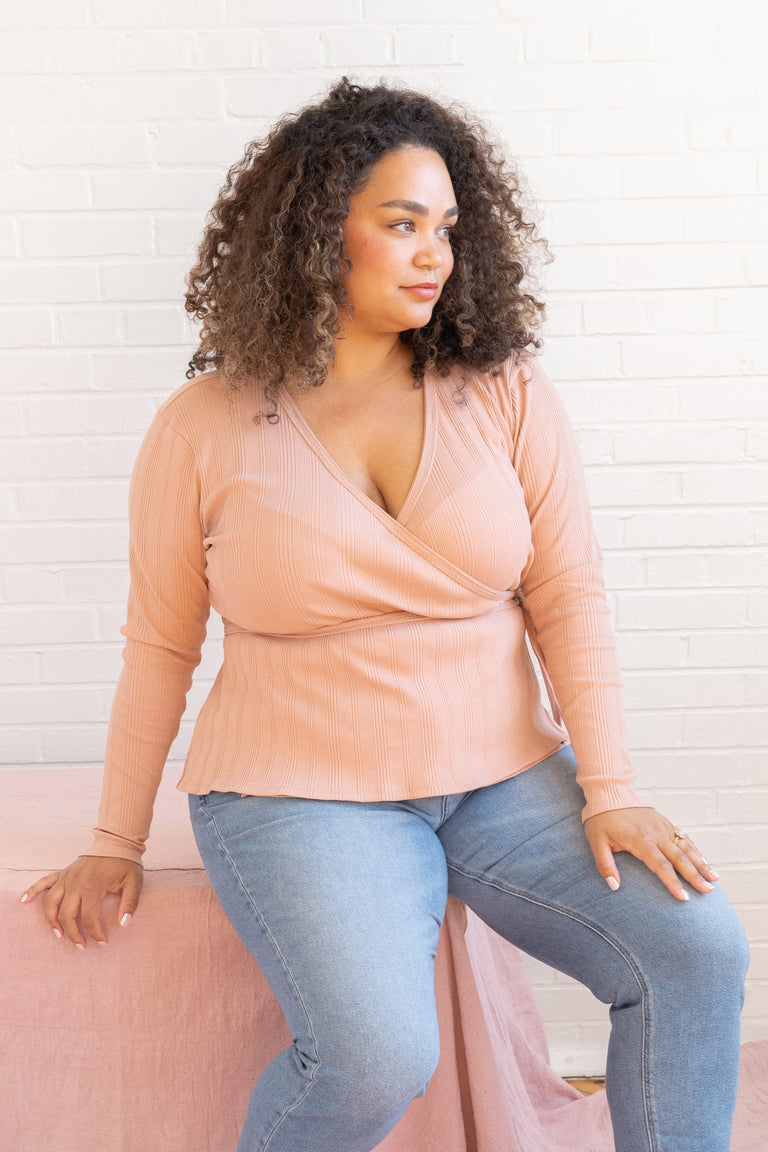 Woman wearing a peach top and blue jeans against a white brick wall.