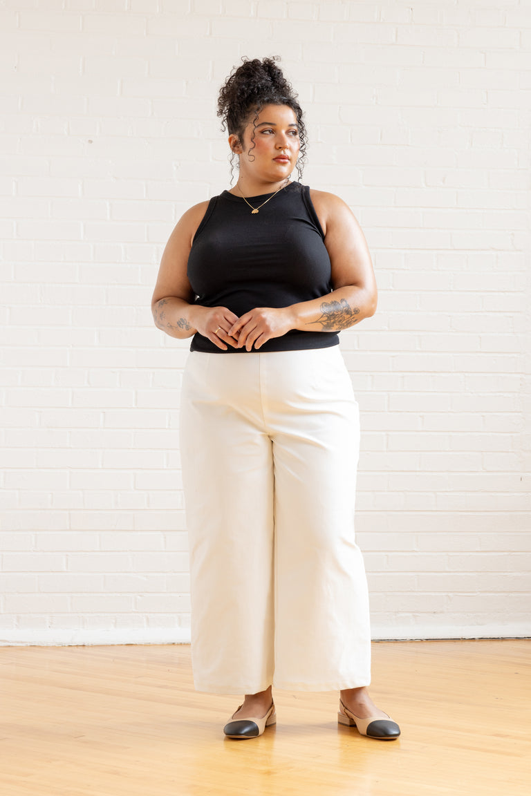 Woman wearing a black sleeveless top and white pants against a white wall.