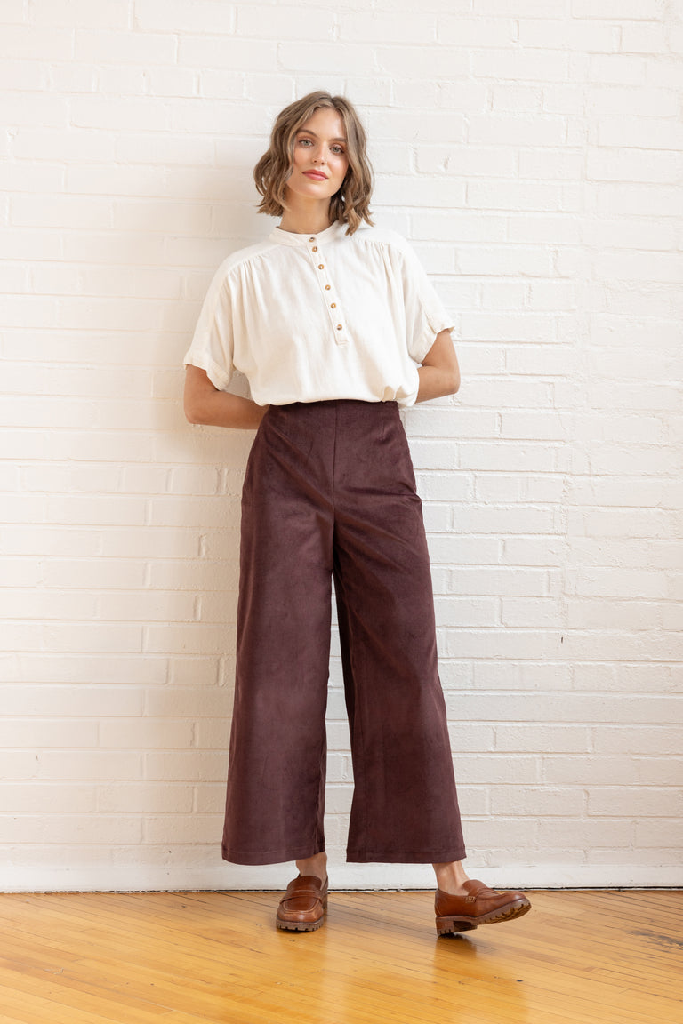 Woman wearing a white blouse and brown pants standing against a white brick wall.