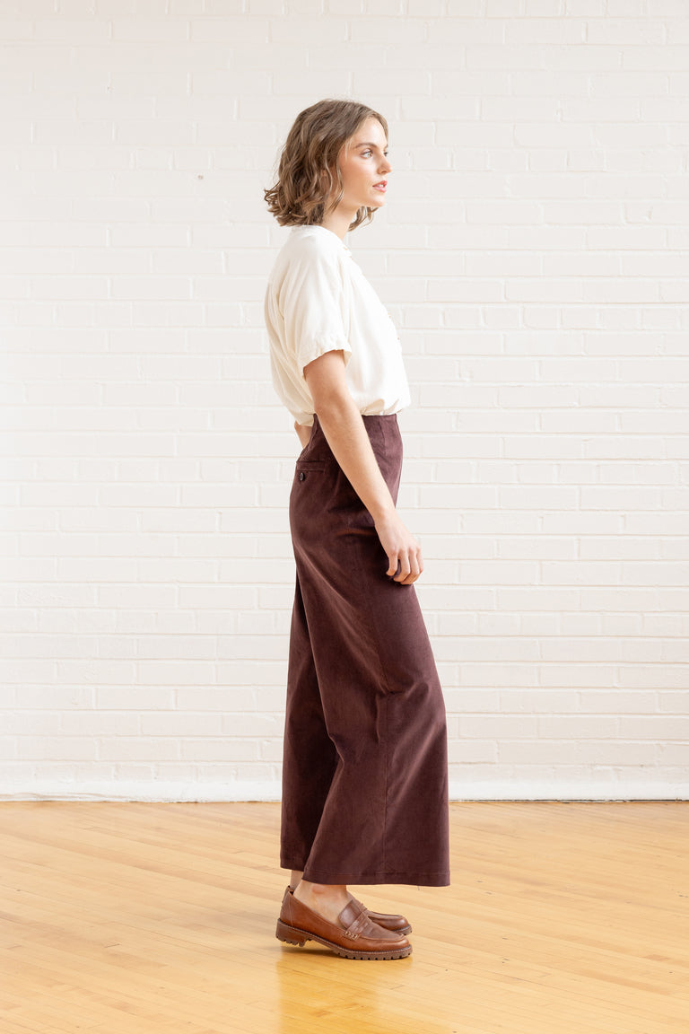Person wearing a white shirt and brown pants standing against a white wall.