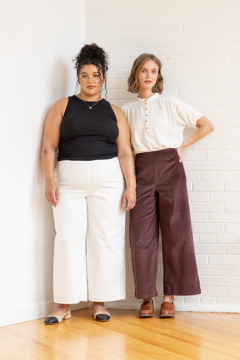 Two women standing against a white brick wall wearing casual outfits.