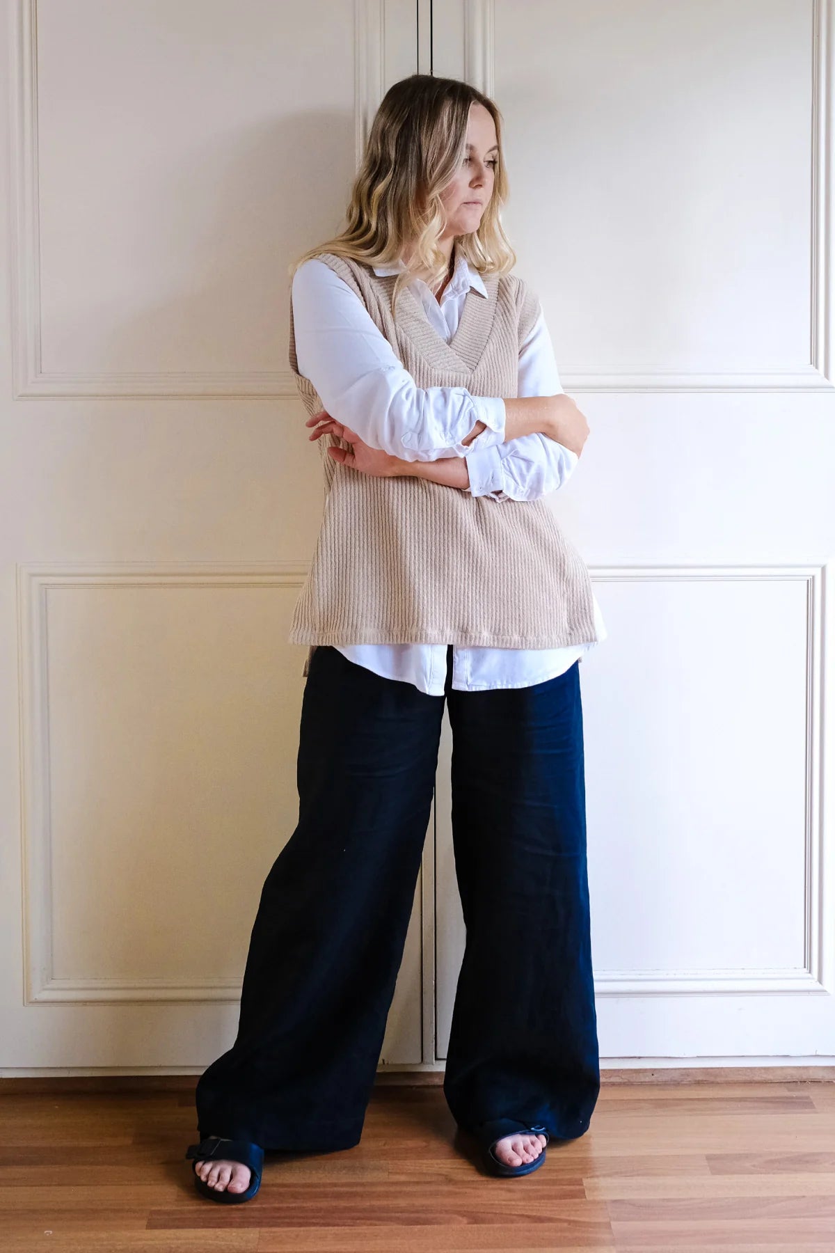 Woman wearing a beige vest over a white shirt with navy pants against a neutral wall.