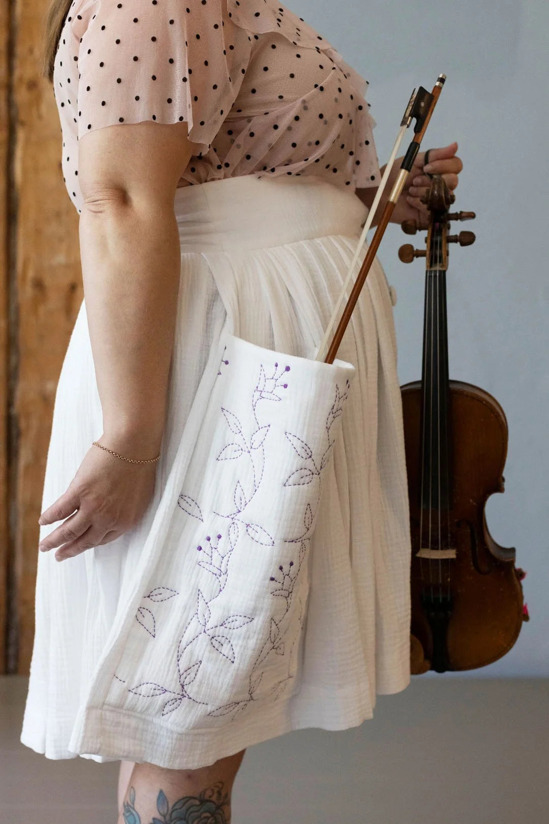 Person holding a violin and a tote bag with floral embroidery against a neutral background