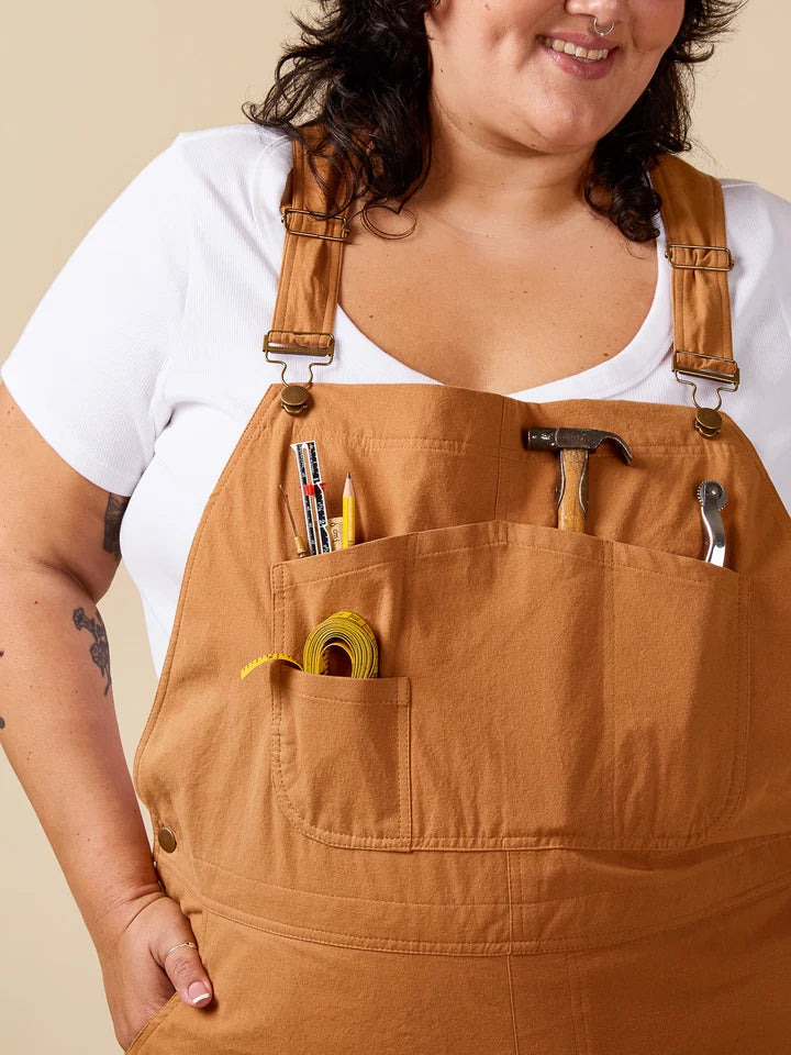 A close-up of a person wearing tan work bib overalls over a white t-shirt. The bib features multiple front pockets containing tools like a hammer, pencil, screwdriver, and tape measure, set against a solid beige background.