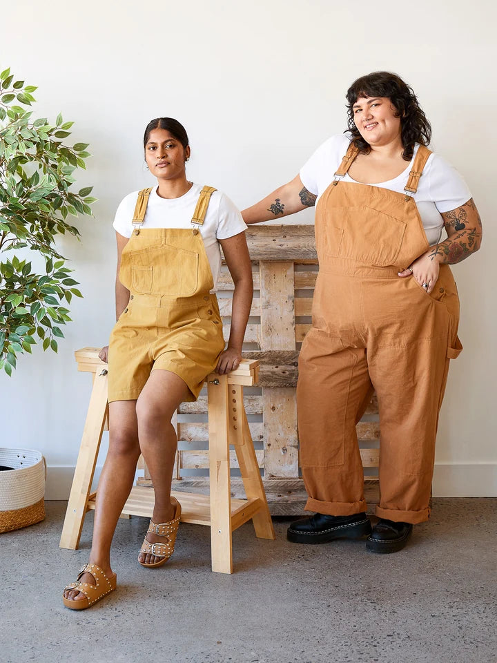 Two women modeling different colorways of linen-look overall jumpsuits. The woman on the left wears a mustard yellow version sitting on a wooden stool, and the woman on the right wears a warm camel brown version standing next to a rustic pallet wood backdrop.