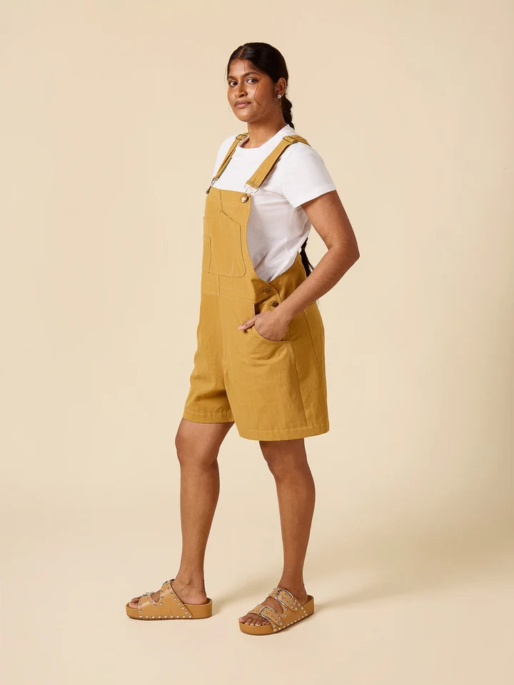 A woman modeling mustard yellow linen bib shorts with adjustable shoulder straps and a white t-shirt underneath, paired with tan woven slide sandals, against a solid beige studio background.
