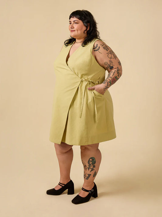 Woman wearing a light green dress with tattoos on her arms and legs against a beige background