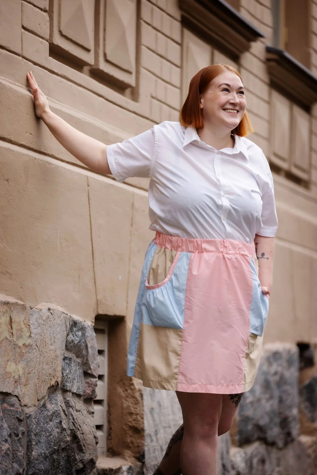 Woman wearing a white shirt and colorful apron standing against a stone wall.