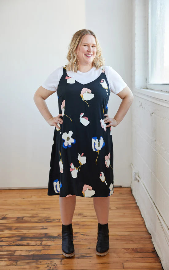 Woman wearing a black floral pinafore dress with a white shirt underneath, standing in a room with wooden flooring and a window.