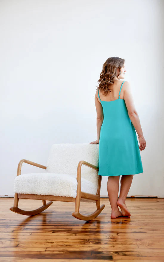 Woman in a teal dress standing next to a wooden rocking chair with a white cushion on a wooden floor.
