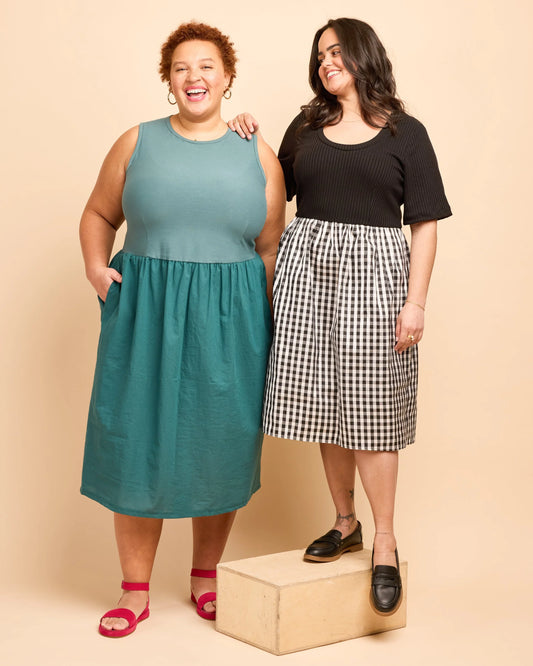 Two women standing together, one in a teal dress and the other in a black top and checkered skirt, on a beige background.