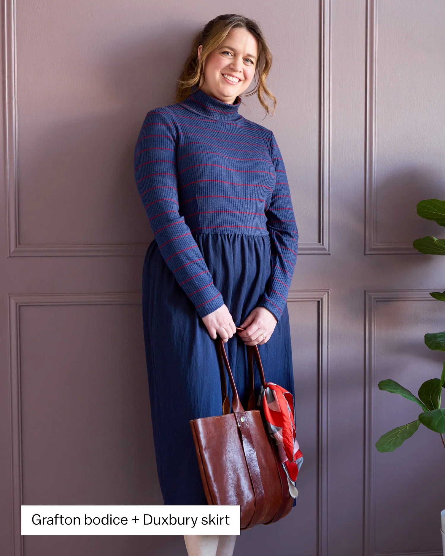 Woman wearing a blue dress holding a brown bag against a purple paneled wall.