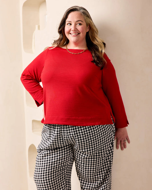 A woman modeling a bright red long-sleeve knit top paired with black and white gingham pants, standing against a beige wall with recessed architectural details.