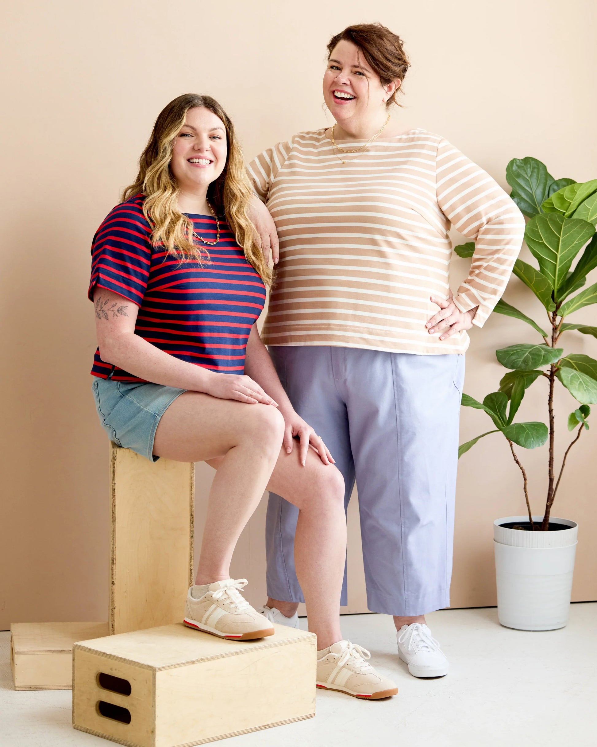 Two women modeling striped long-sleeve tops in a studio setting. One woman sits on a wooden box wearing a navy and red striped shirt with denim shorts, and the other stands wearing a beige and white striped shirt with light blue cropped pants.