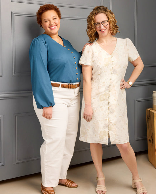 Two women standing against a gray paneled wall, one in a blue top and white pants, the other in a white dress.