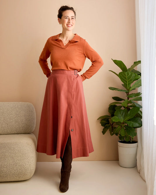 Woman wearing a rust-colored top and skirt standing in a room with a plant and chair.