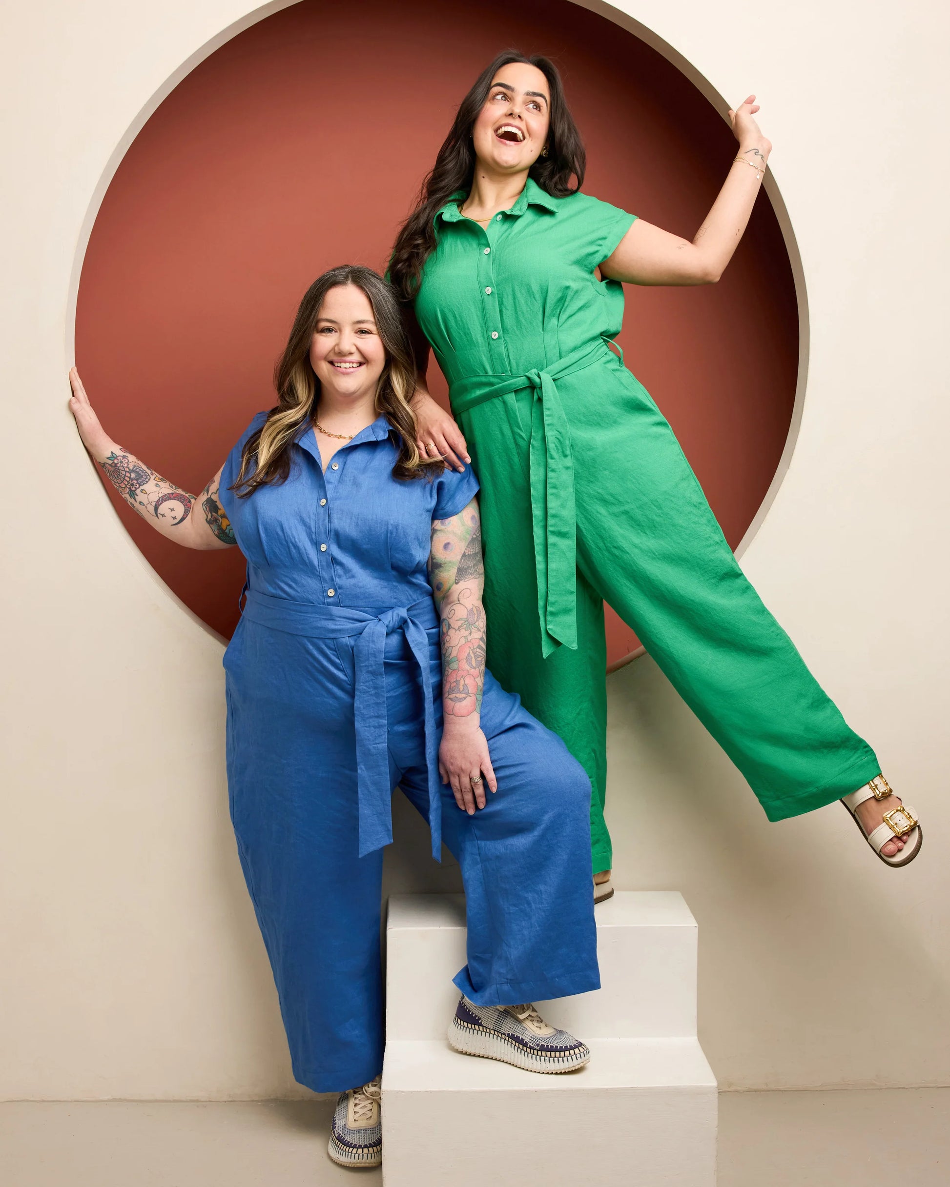 Two women in colorful jumpsuits posing against a beige wall with a circular cutout.