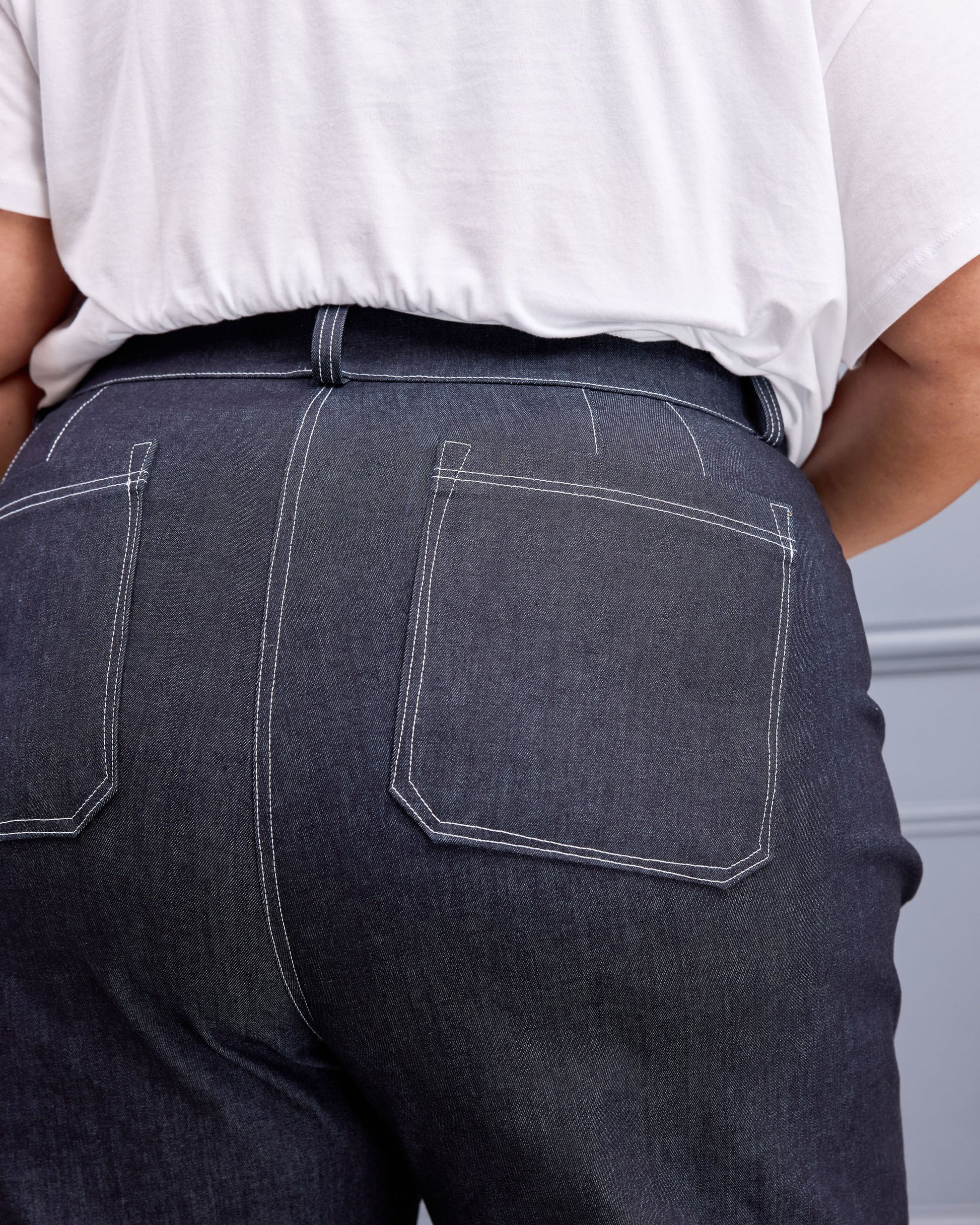 Close-up back view of a person wearing dark indigo denim jeans with white contrast stitching and a white t-shirt.