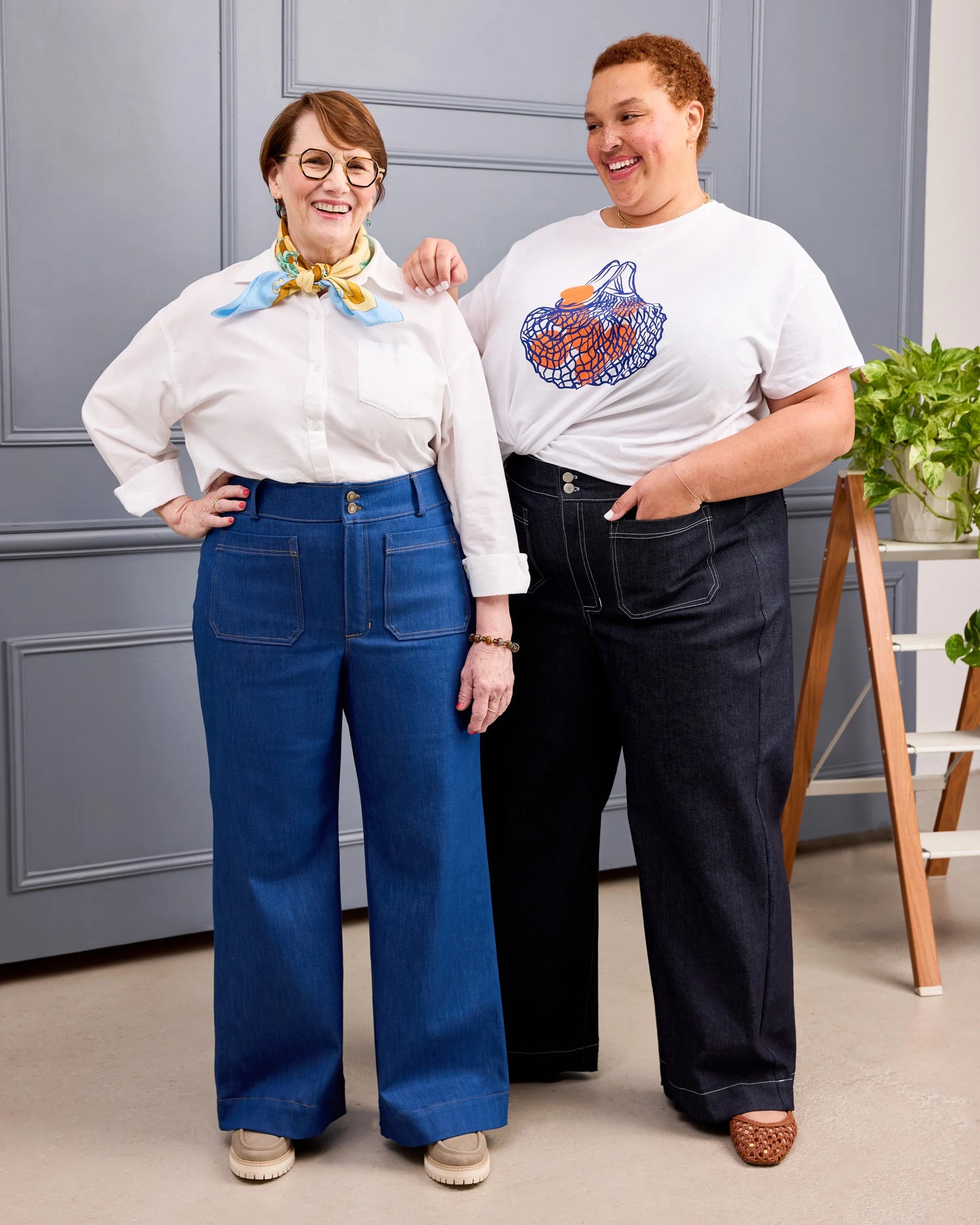 Two women modeling wide-leg jeans in a studio setting. The woman on the left wears a white blouse with a blue and yellow neck scarf and blue jeans.