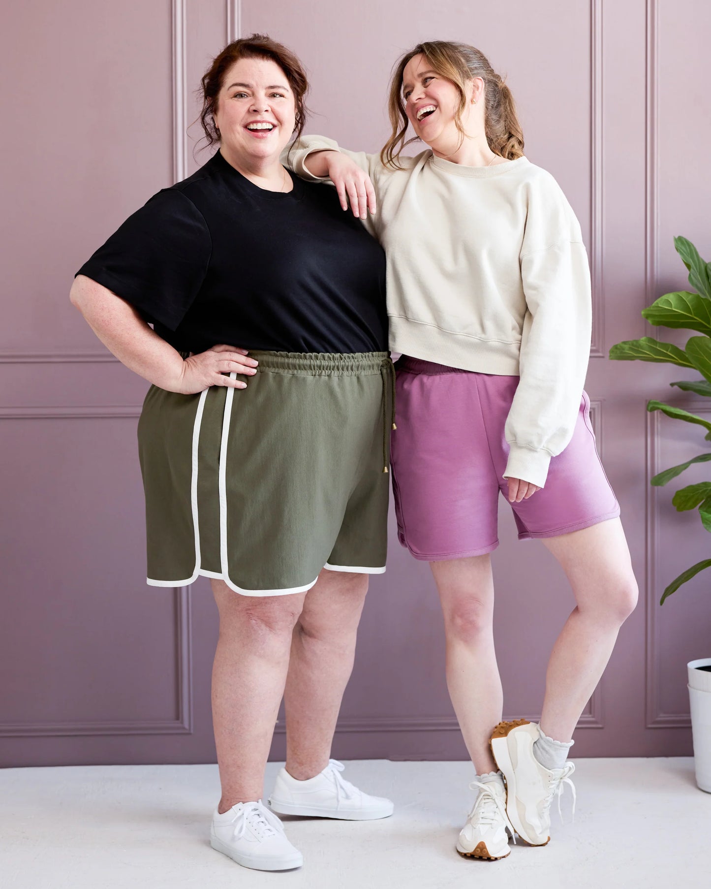 Two women standing together wearing athletic wear against a light purple wall.
