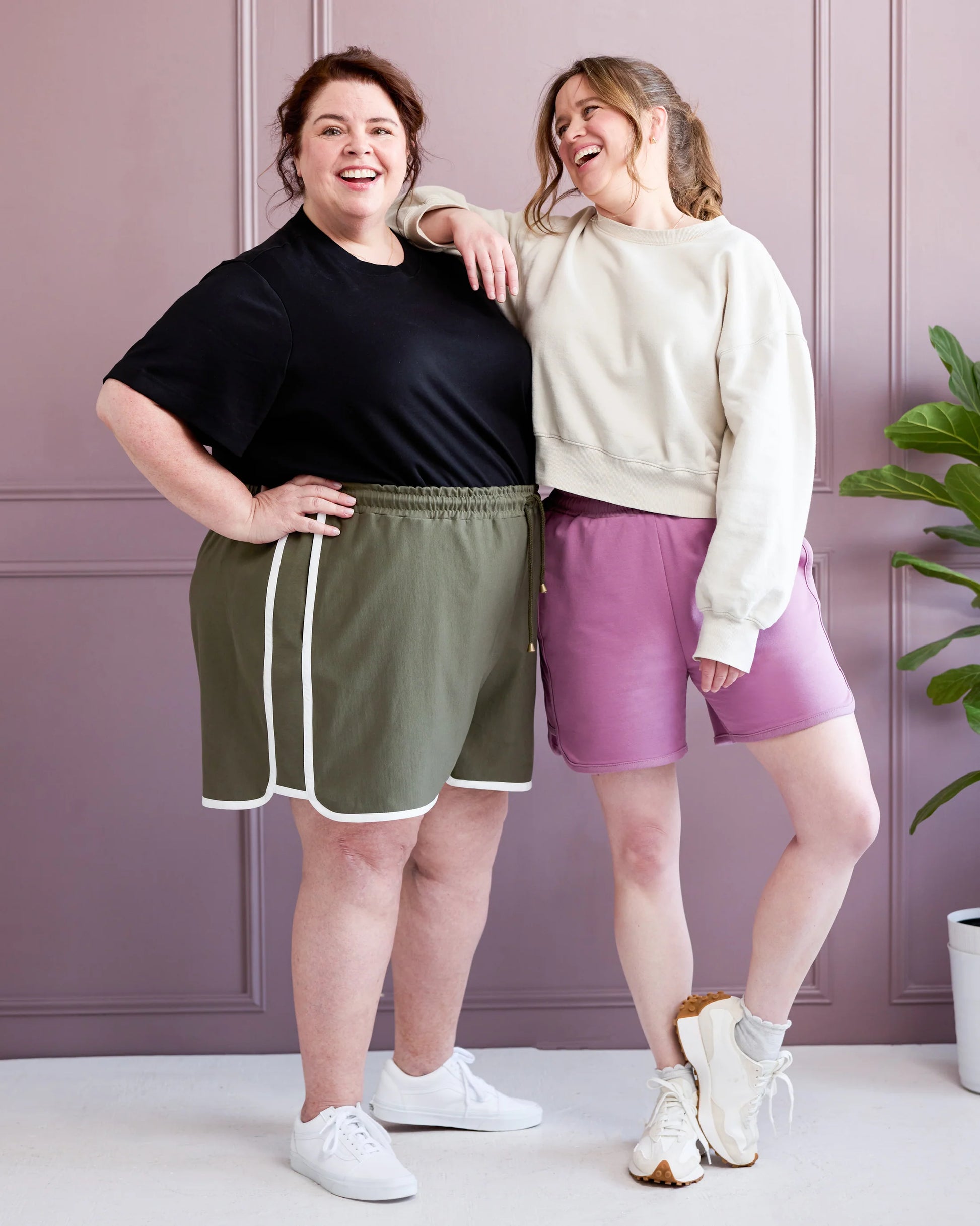 Two women standing together wearing athletic wear against a light purple wall.