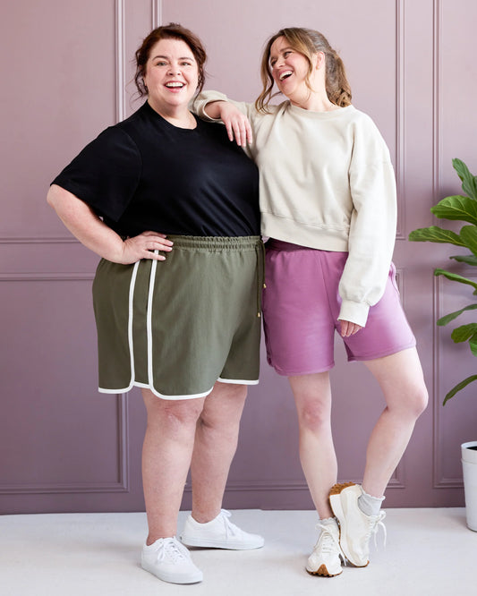 Two women standing together wearing athletic wear against a light purple wall.