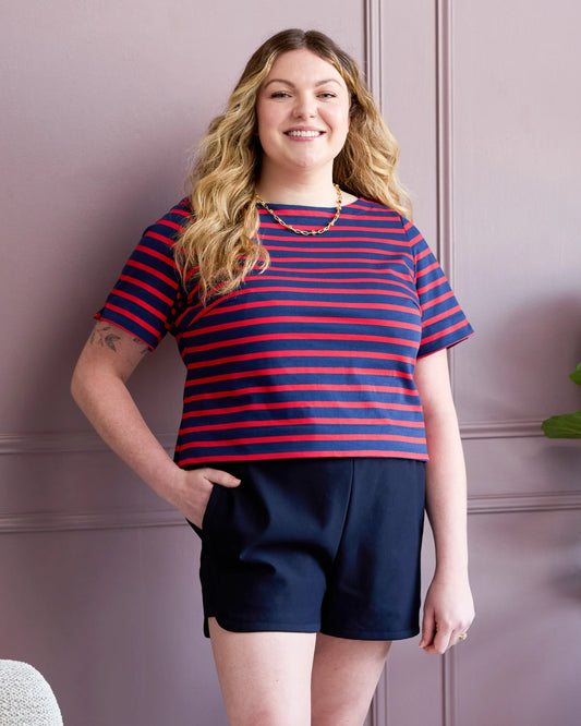Woman wearing a red and blue striped shirt and navy shorts against a pink wall.