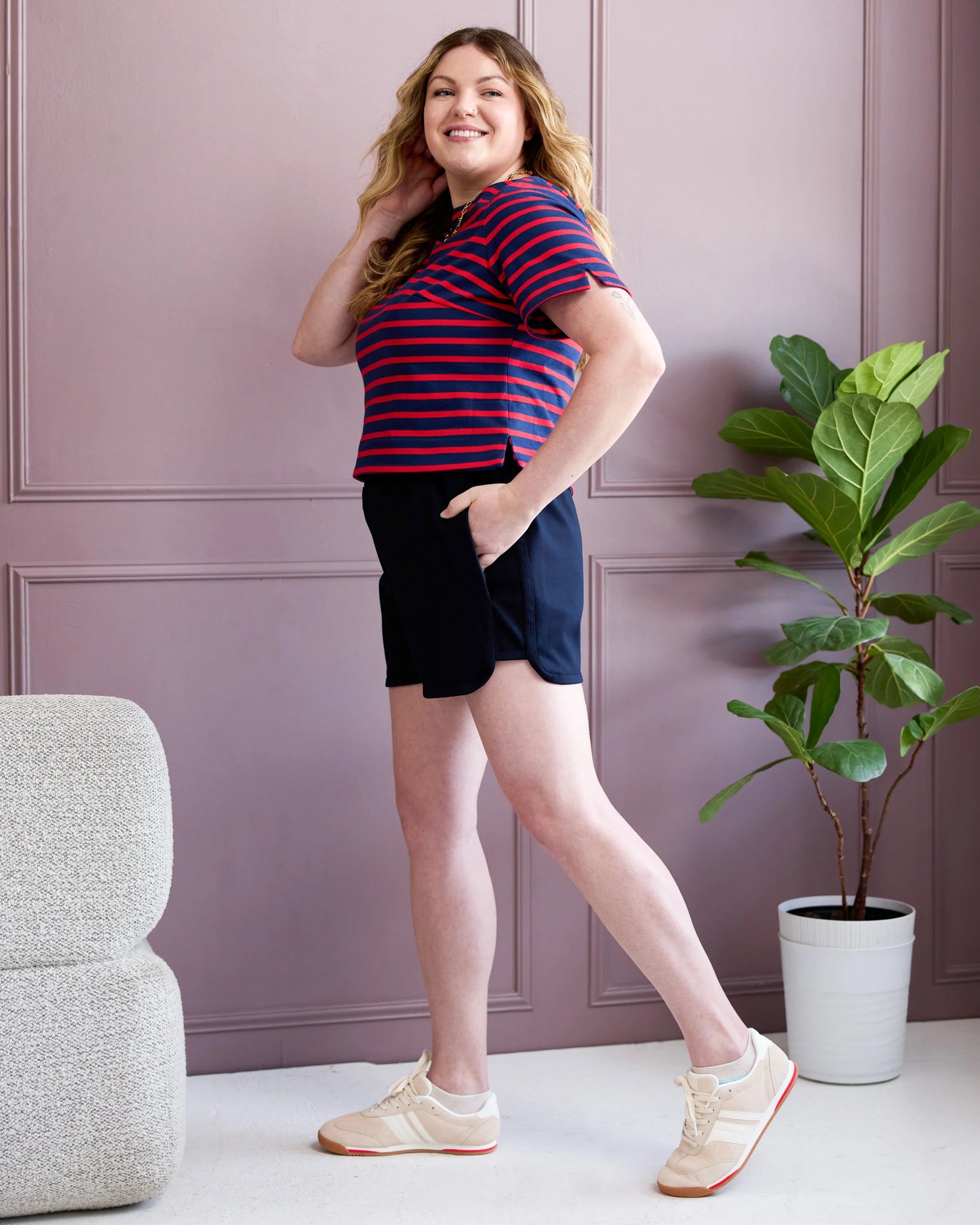 Woman in a striped shirt and shorts standing in a room with a plant and chair.