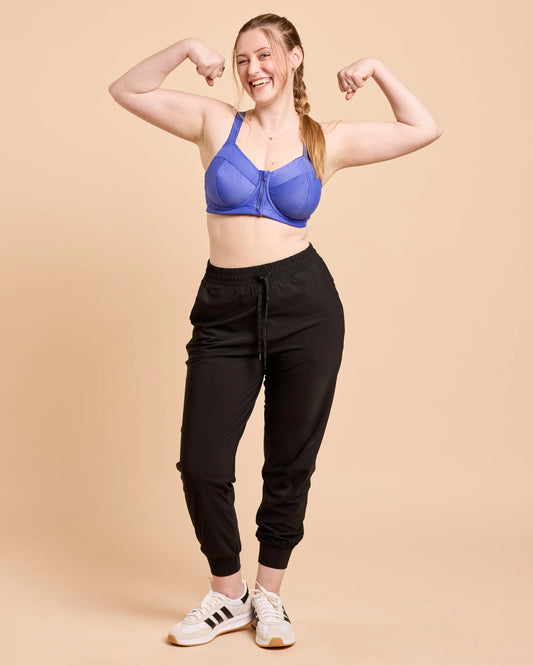 Woman flexing her muscles wearing a blue sports bra and black jogger pants on a beige background