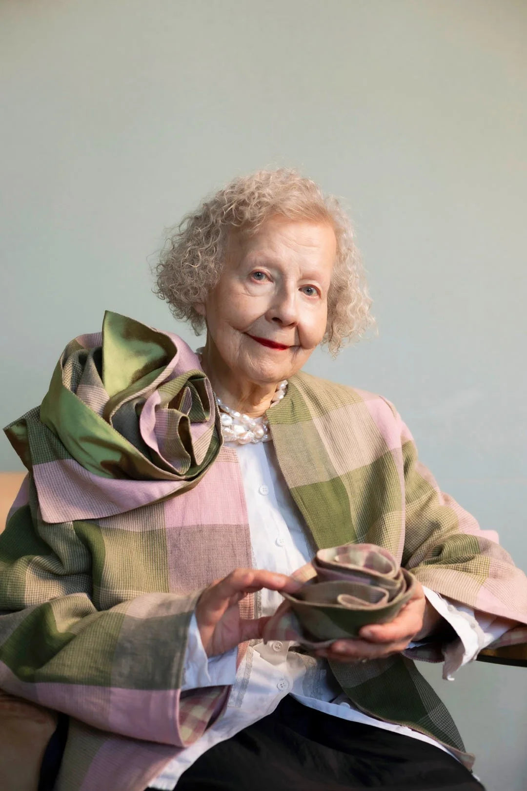 Woman holding a small round object with a checkered shawl over her shoulders against a plain background