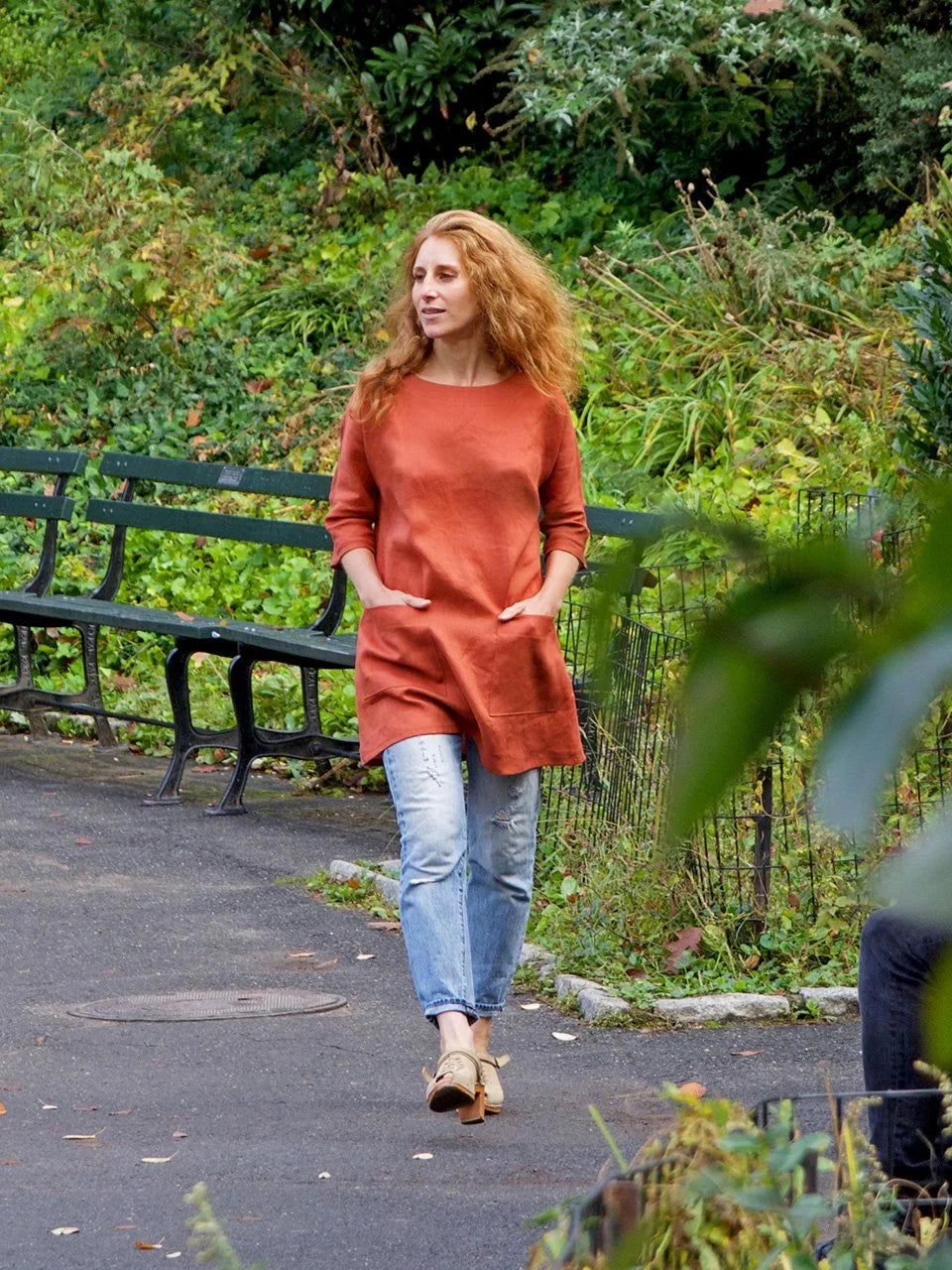Woman walking in a park wearing a rust-colored tunic and blue jeans.
