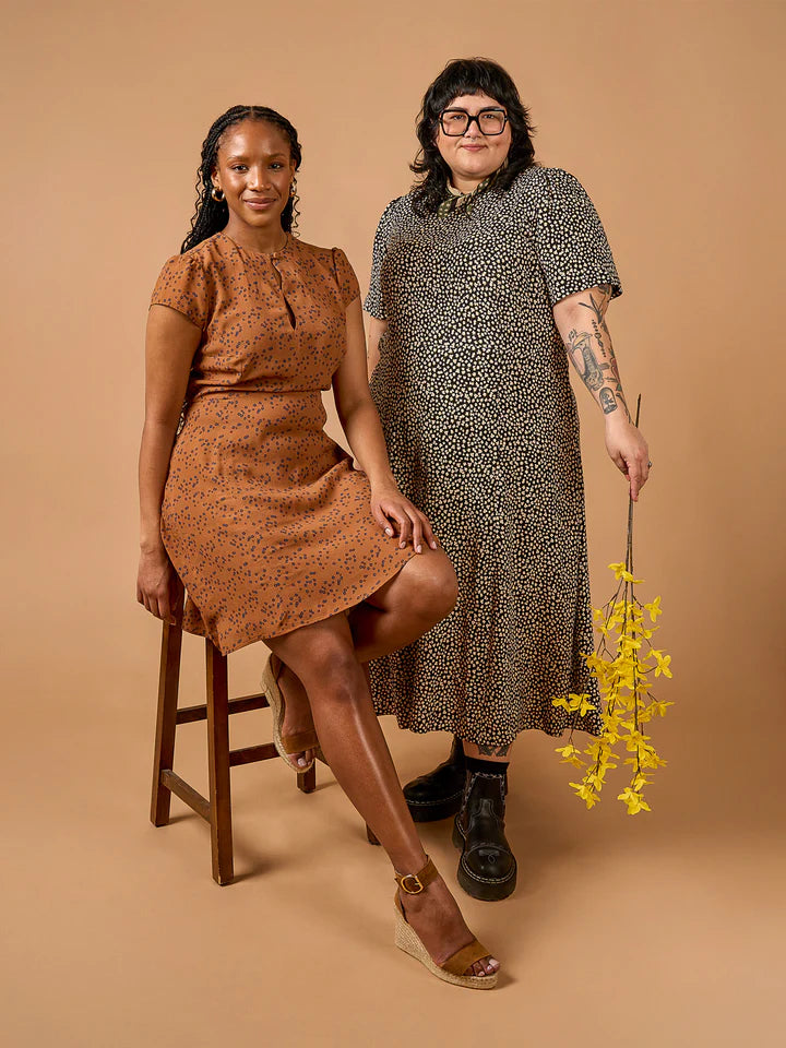 Two women posing together against a beige background, one on a stool holding a plant.