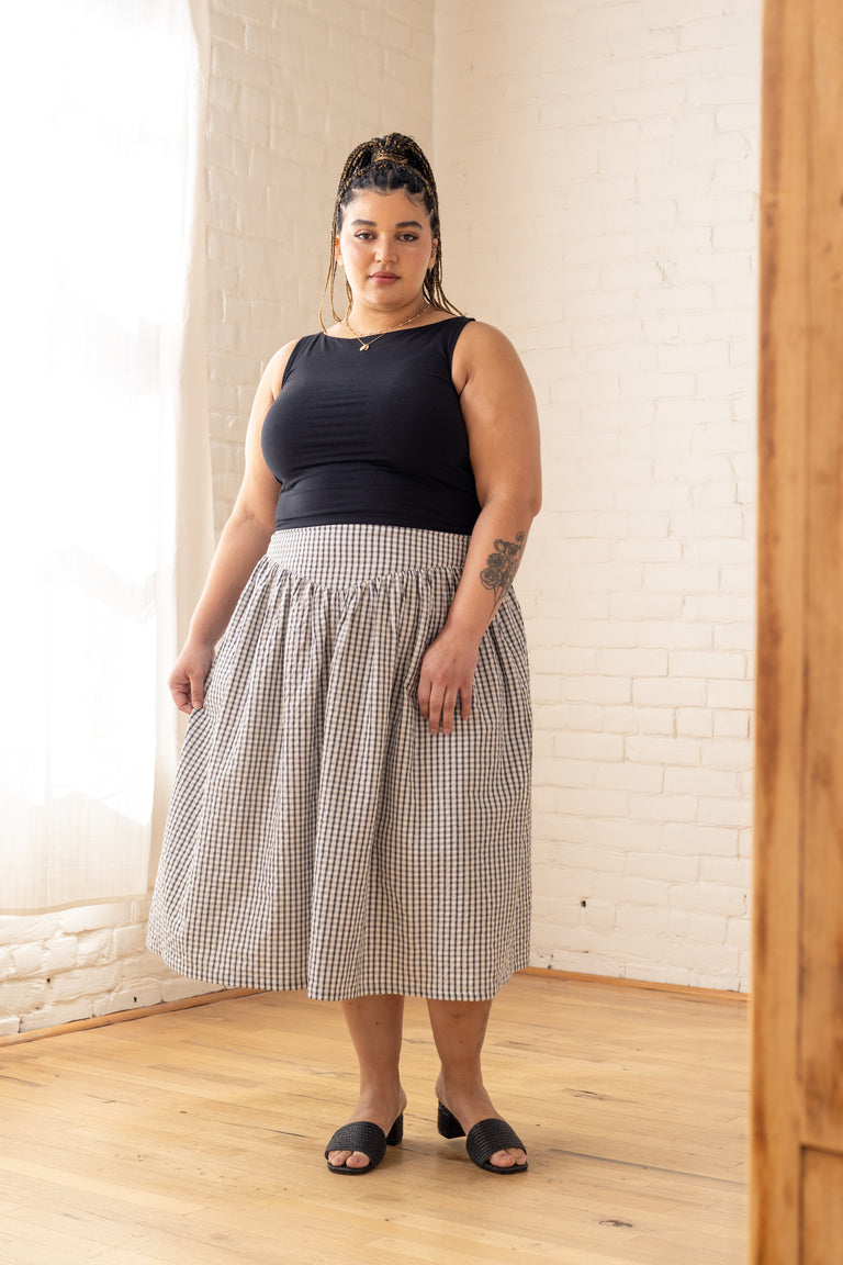 Woman wearing a black top and checkered skirt standing in a room with white brick walls.