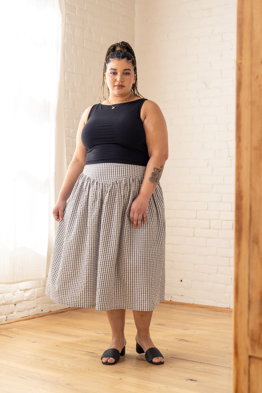 Woman wearing a black top and checkered skirt standing in a room with white brick walls.