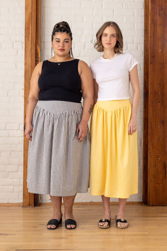 Two women standing side by side wearing a black sleeveless top and checkered skirt, and a white t-shirt and yellow skirt, against a white brick wall.