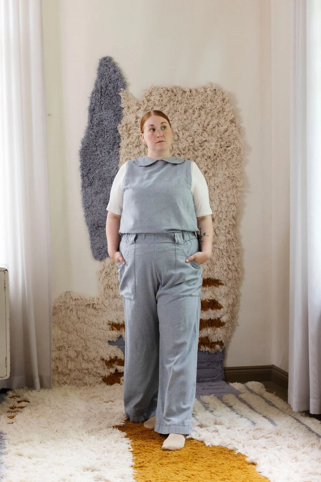 Woman in a gray outfit standing in front of textured wall art in a room with curtains.